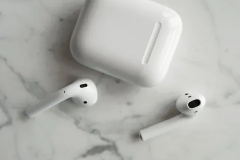 Seseorang sedang memegang sepasang earphone tanpa kabel berwarna putih di samping wadah pengisi daya